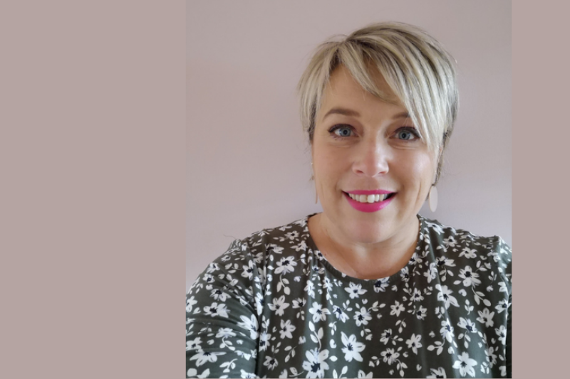 White female with blonder hair waring a green floral print top standing in front of a plain mauve background